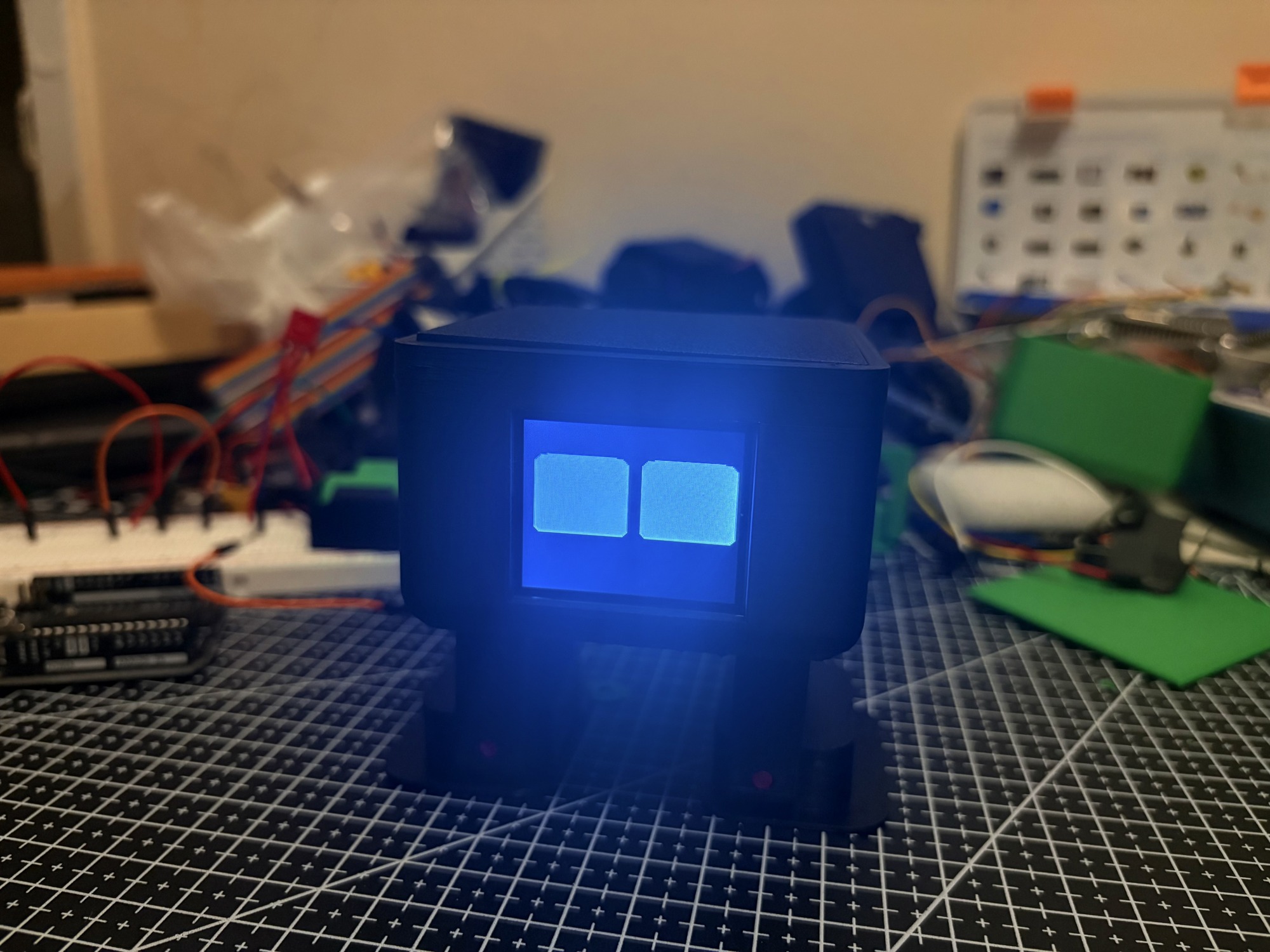 LUMA, a small blue 3D printed robot with two glowing eyes, sitting on a cutting mat
